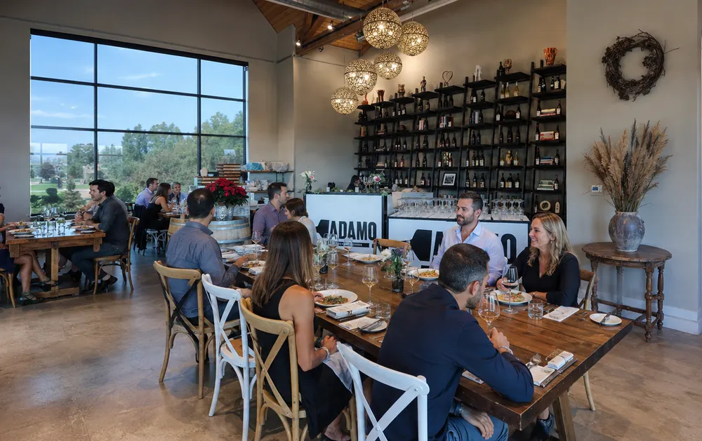Diners at long shared tables in the Adamo Estate dining room, with a floor-to-ceiling window looking out over the property.