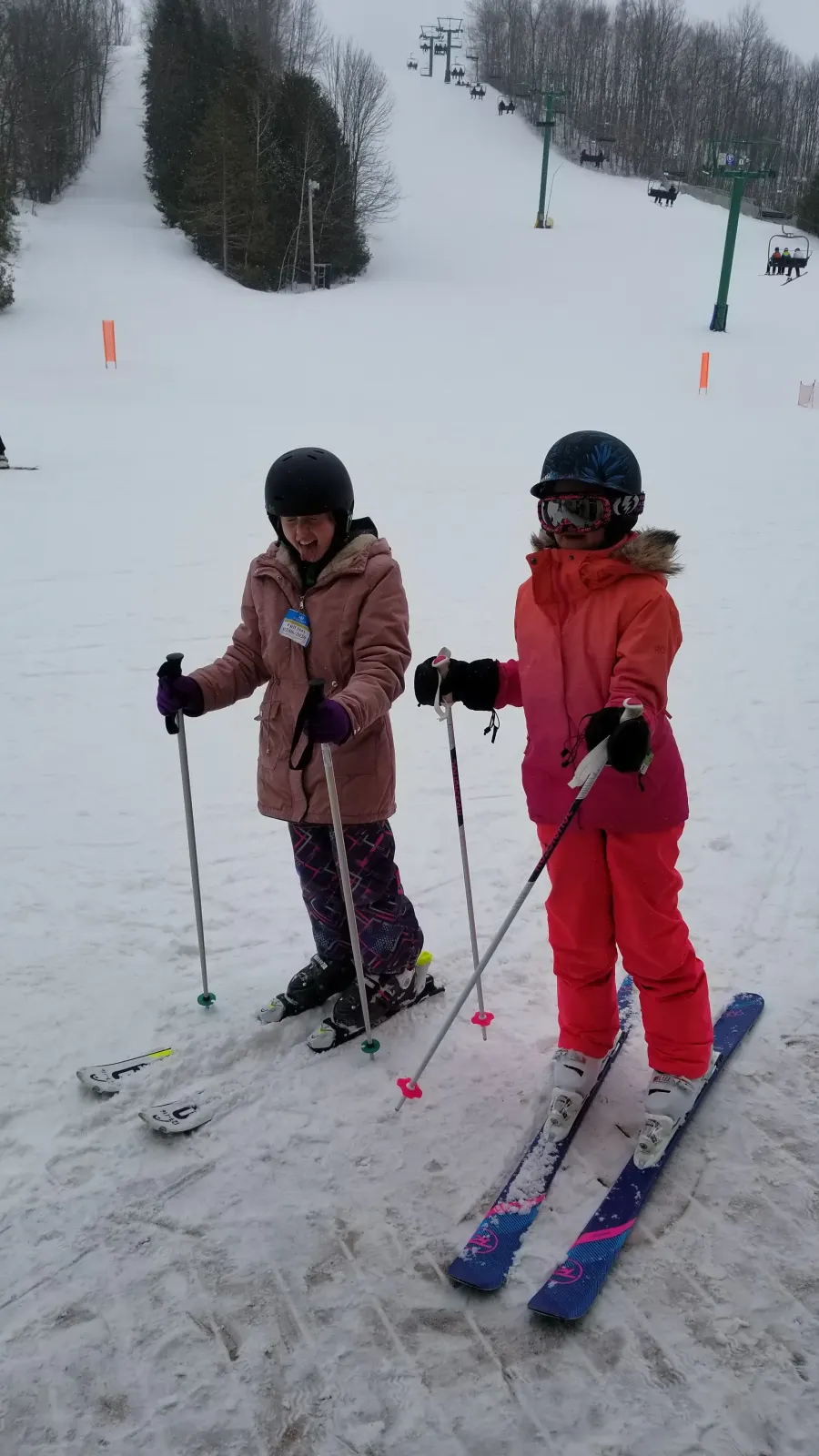 Two young skiers with poles and lift tickets standing on the snow at the base of the slope, chairlift climbing the hill behind them.