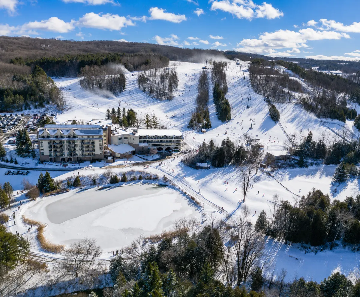 Aerial view of the Hockley Valley ski hill on a busy winter day, with skiers spread across every run, snow guns firing along the ridge and the frozen pond below the hotel.