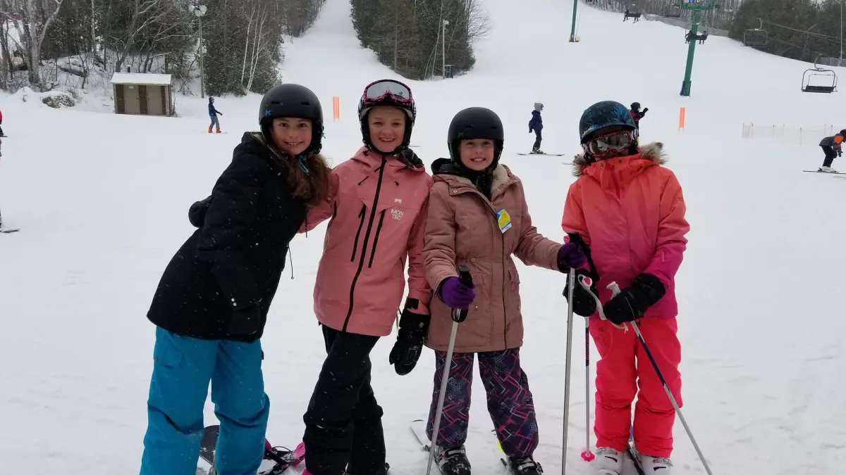 Four young skiers in helmets and winter gear standing together at the bottom of the hill, with the runs and chairlift behind them.