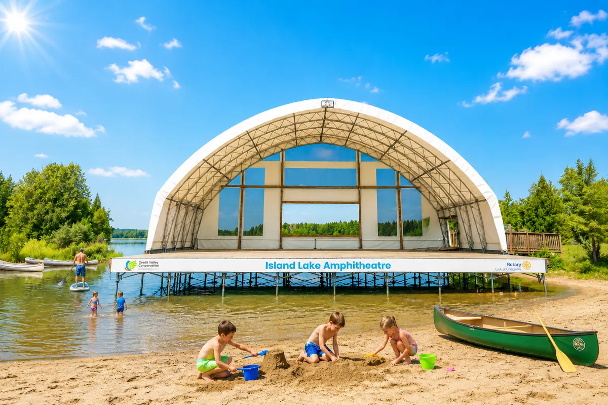 The Island Lake Amphitheatre stage built out over the water, with children building sandcastles on the beach in front and a paddleboarder passing behind.