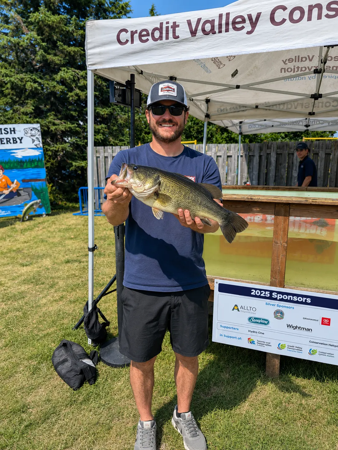 An angler holding a large largemouth bass in front of the Credit Valley Conservation tent at the Island Lake Bass Derby, with the sponsor board visible behind him.