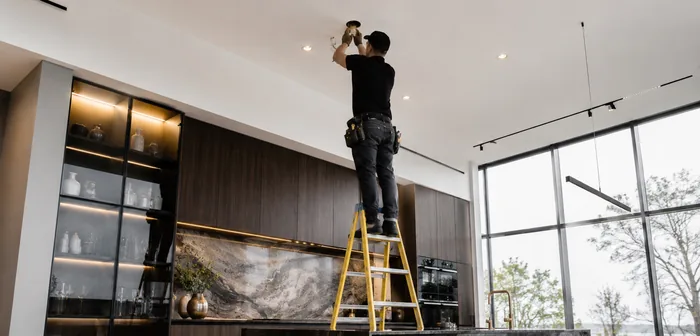 An electrician on a step ladder fitting a pot light into the ceiling of a modern kitchen.