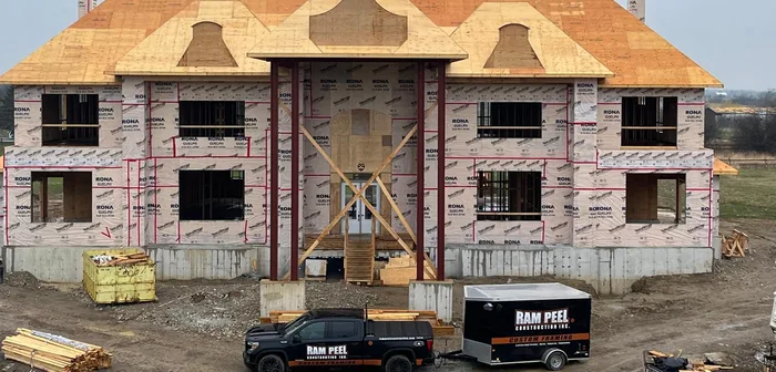 A large custom home framed and sheathed to the roof deck, with a Ram Peel Construction pickup and equipment trailer parked out front.