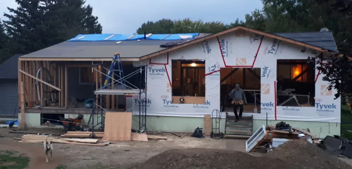 A single-storey home addition wrapped in Tyvek at dusk, roof decked and scaffolding still standing.