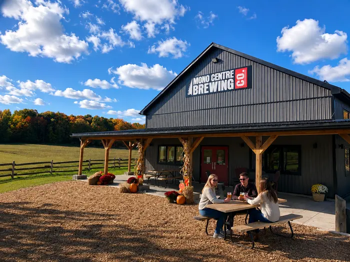 The dark barn-style Mono Centre Brewing Co. building on an autumn afternoon, with people drinking at a picnic table out front and pumpkins and cornstalks by the door.