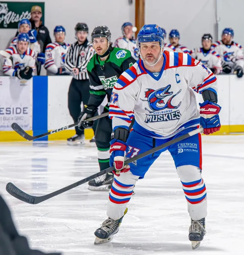 A Shelburne Muskies player wearing the captain's C skating up ice, with the team bench and an opposing player behind him.