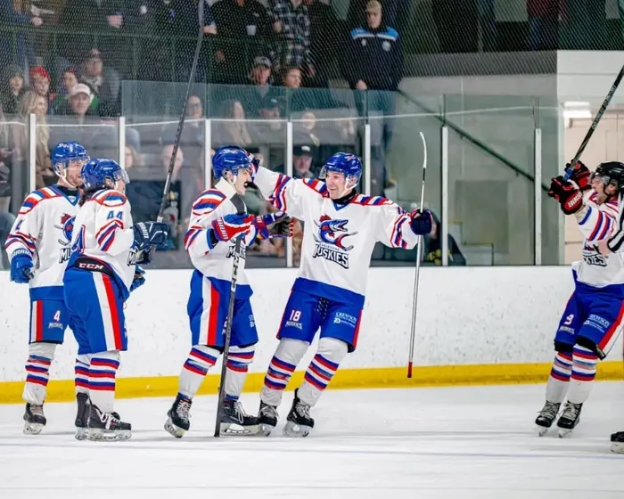 Shelburne Muskies players in red, white and blue jerseys celebrating a goal in front of a full crowd at the Centre Dufferin Recreation Complex.