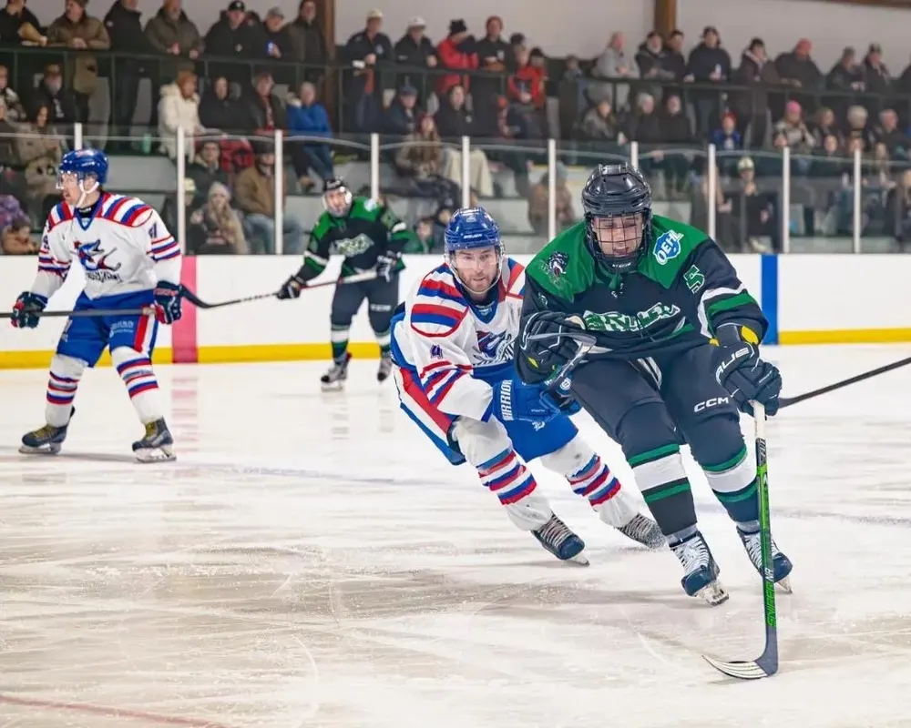 A Muskies player racing an opposing forward for the puck with a packed grandstand of spectators behind the glass.