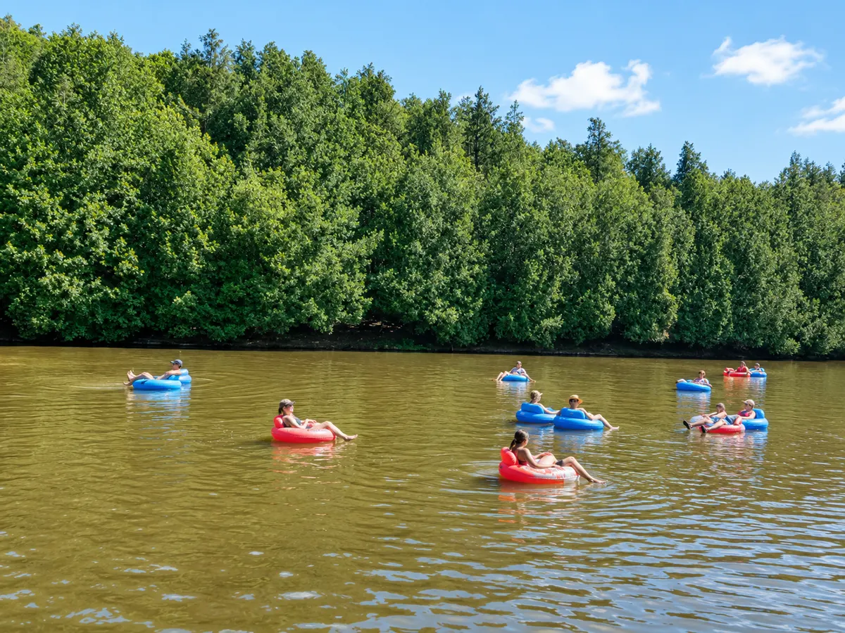 A dozen people floating on inflatable tubes down a wide, calm stretch of the Grand River, with dense green forest along the far bank.